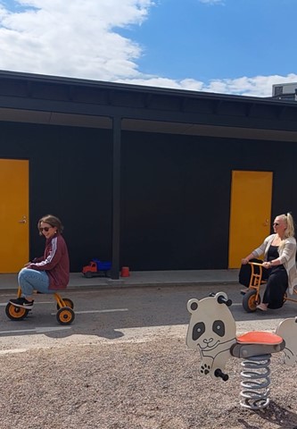 Kaksi henkilöä päiväkodin pihalla, ajavat lasten polkuneuvoilla ja nauravat.