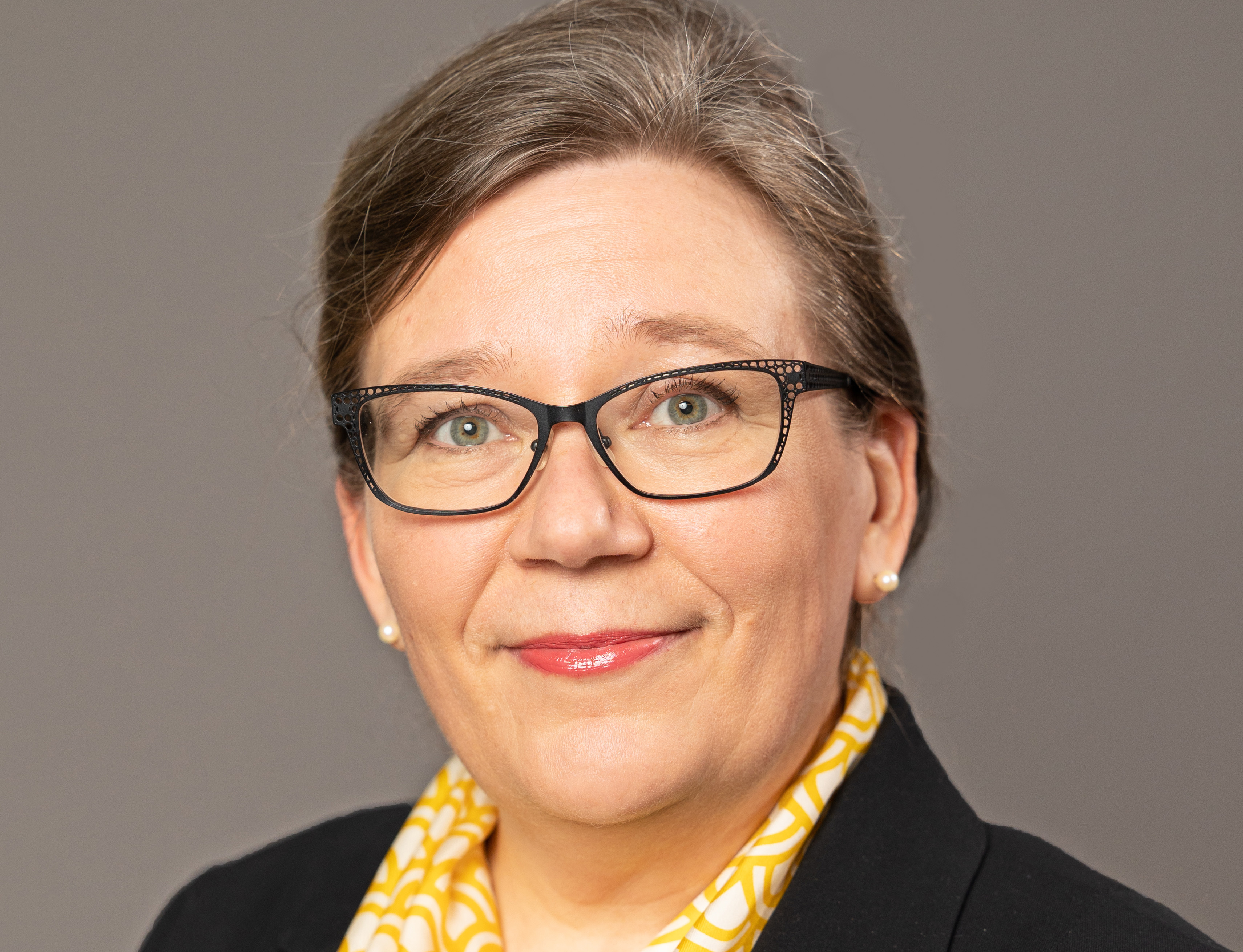 Kuvassa nainen, jolla on silmälasit ja hymyilevä ilme, pukeutuneena mustaan jakkuun ja keltaiseen kuviolliseen huiviin. Tausta on neutraali harmaa.