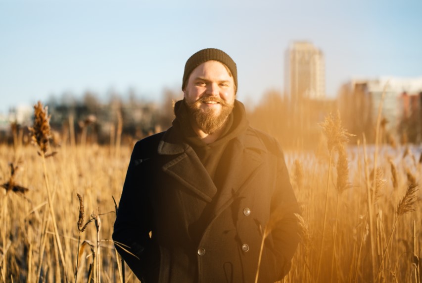 Mies seisoo ulkona viljapellossa. Hänellä on päällään pipo ja takki.