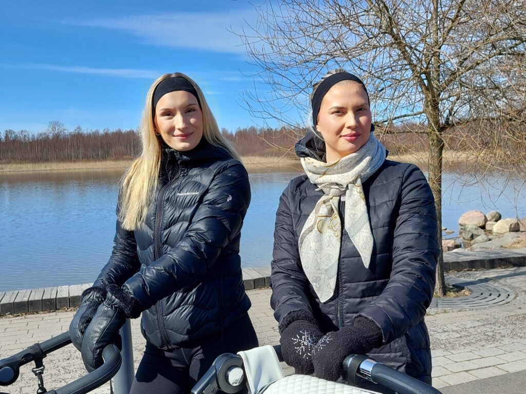 Kaksi nuorta naista seisovat rantapuistossa lastenvaunujen kanssa, takana järvinäkymää.