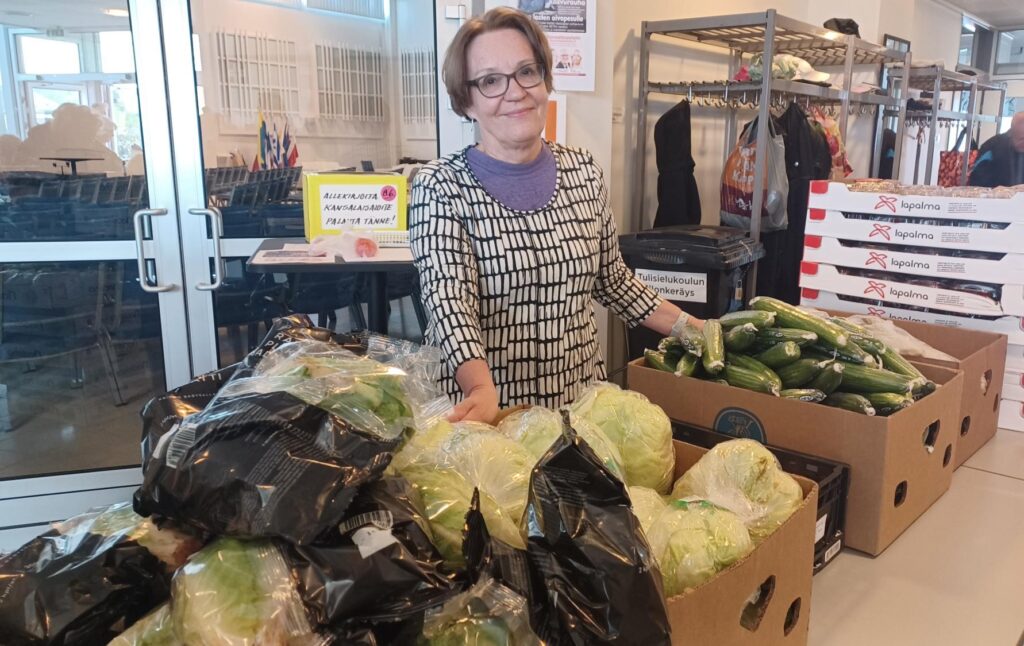 Nainen jakamassa vihanneksia. Laatikoihin on pakattuna kurkkua, kaalinkeriä ja salaattia.
