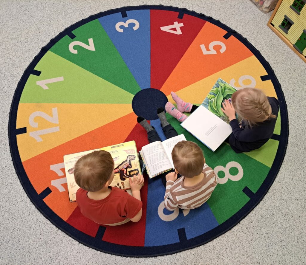 Kolme lasta lukemassa kirjoja istuen värikkäällä matolla, jossa on numeroita. Kuva on otettu ylhäältäpäin.
