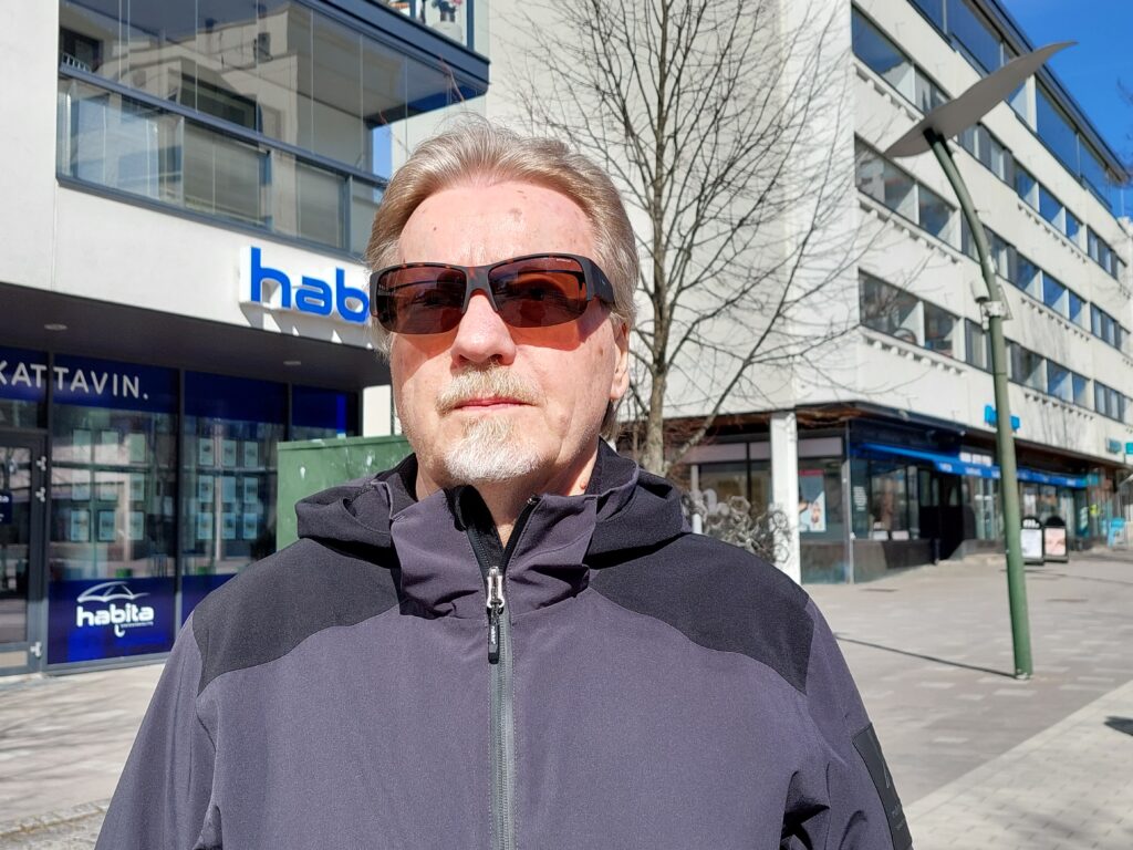 Mies lähikuvassa, hänellä on aurinkolasit päässä. Taustalla kerrostalonäkymää.
