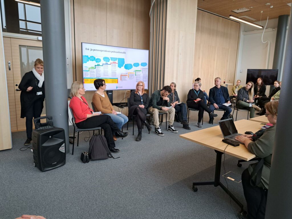 Useampi henkilö istuu digitaalisen esityksen edessä, paneelikeskustelu alkamassa. 