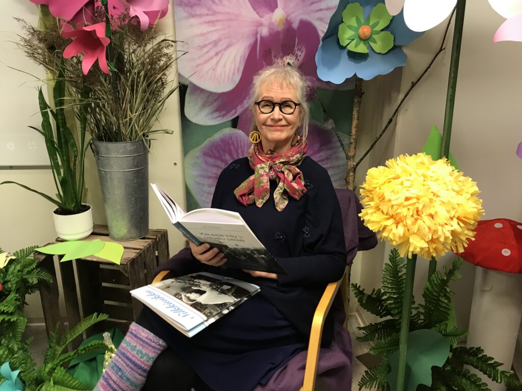 Nainen istumassa tuolissa, hänellä on kädessä kirja ja sylissä lehtiä. Ympärillä on värikkäitä kukkakoristeluja.