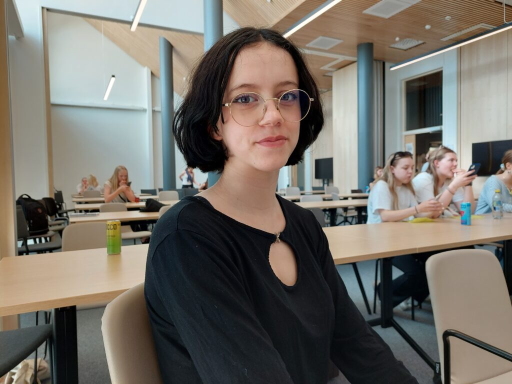 Nuori henkilö istumassa kokoushuoneessa. Hänellä on musta paita yllään. Takana näkyy muita henkilöitä.
