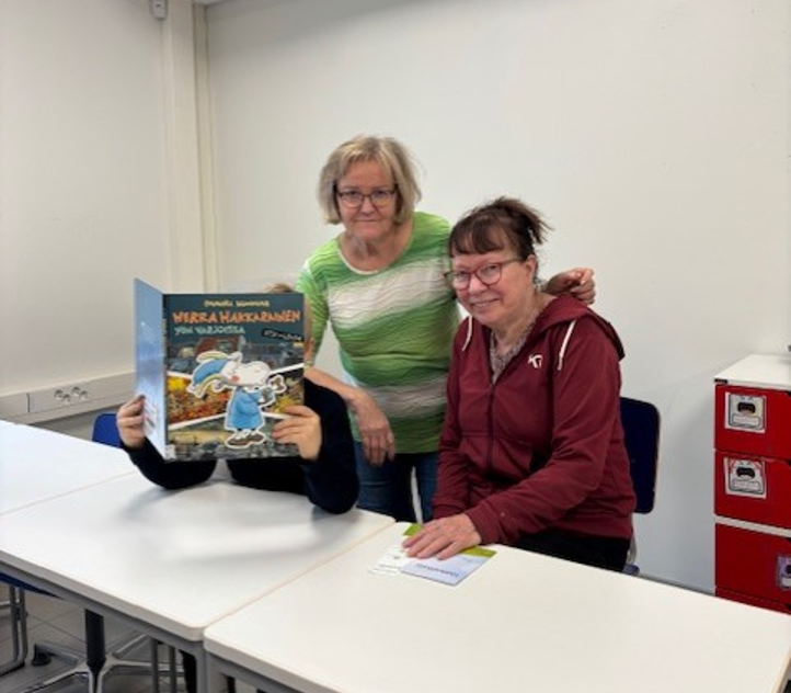 Kaksi naista ja lapsi istuvat pöydän takana. Lapsi lukee Herra Hakkarainen -kirjaa.