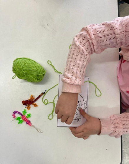 Ylhäältä päin otettu kuva, jossa näkyy lapsen kädet. Lapsi tekee ompelutyötä paperiin. Vieressä on vihreä lankakerä.