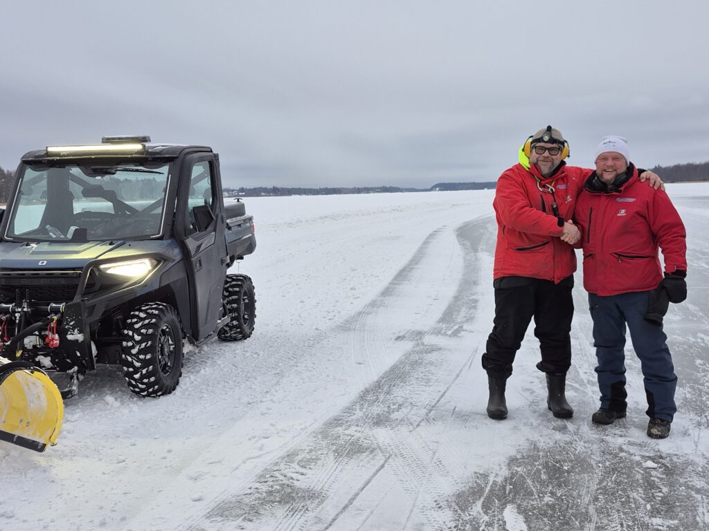 Kuvan vasemmassa reunassa on koppimönkijä, jossa aura edessä. Jäällä seisoo kaksi punatakkista miestä, jotka kättelevät toisiaan ja hymyilevät.