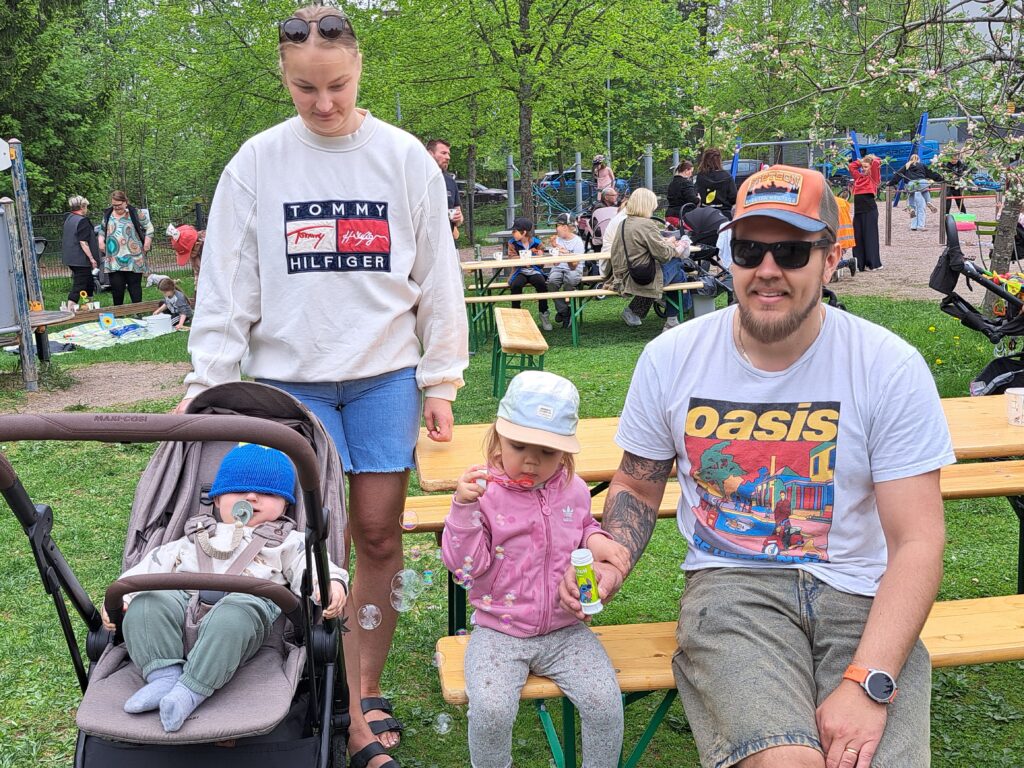 Äiti ja isä kahden pienen lapsen kanssa. Vauva on rattaissa ja isompi lapsi puhaltelee saippuakuplia. Taustalla ihmisiä.