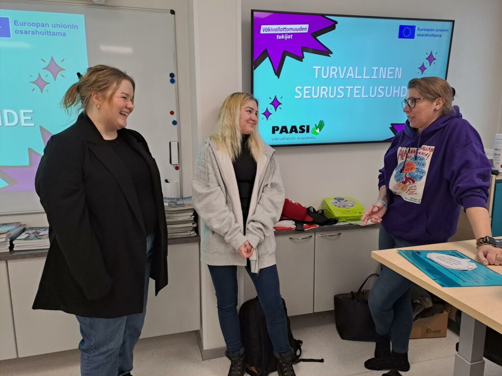 Kolme henkilöä luokkahuoneessa, taustalla esitysmateriaalia otsikolla "Turvallinen seurustelusuhde".