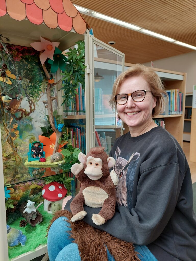 Naisella on apinapehmoeläin sylissä ja taustalla näkyy vitriini, jossa on satumaisemaa esillä.