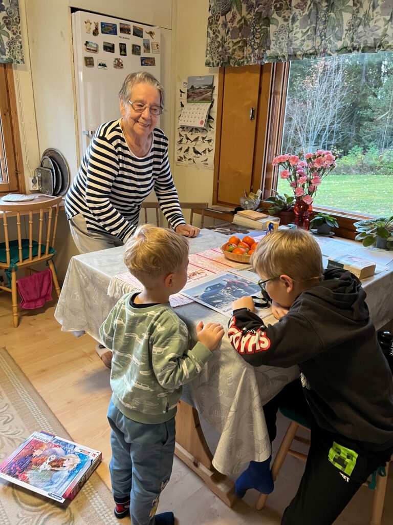 Vanhempi nainen ja kaksi lasta keittiössä ikkunan ääressä. Pojat selailevat sanomalehteä pöydän ääressä.