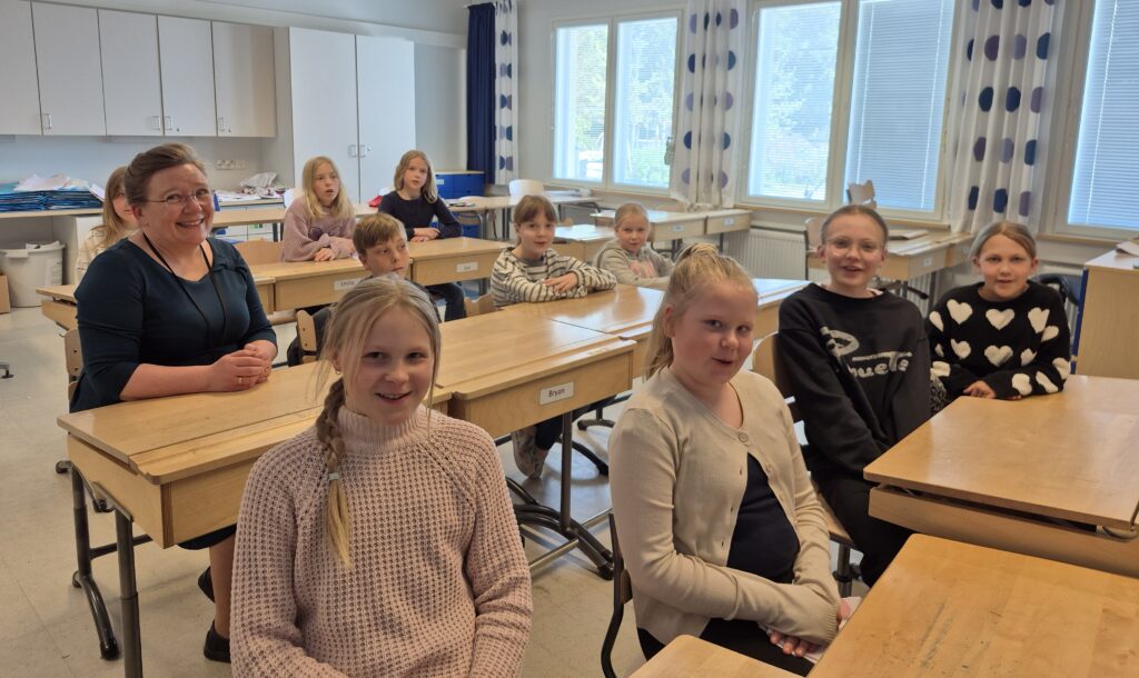 Oppilaat ja yksi aikuinen henkilö istuvat pulpeteissa luokkahuoneessa. He näyttävät iloisilta.