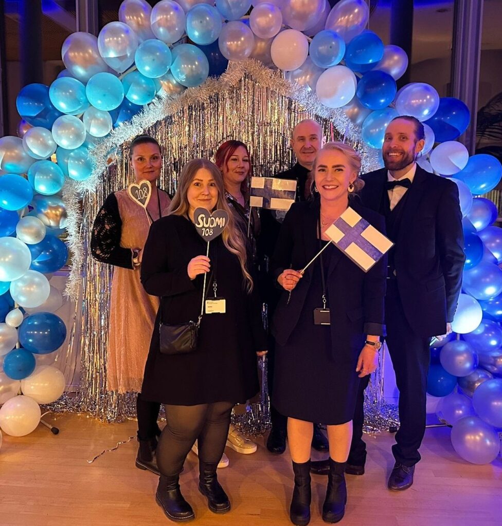 Ihmisiä seisoo Suomi-liput ja koristeet kädessään. Taustalla on koristeltu seinä, jossa on sini-valkoisia ilmapalloja.