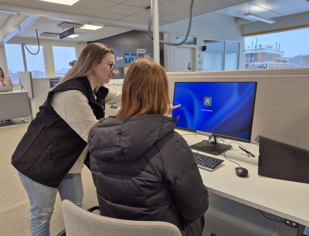 Kaksi henkilöä on yleisötietokoneen äärellä. Toinen henkilöstö opastaa istumassa olevaa henkilöä.