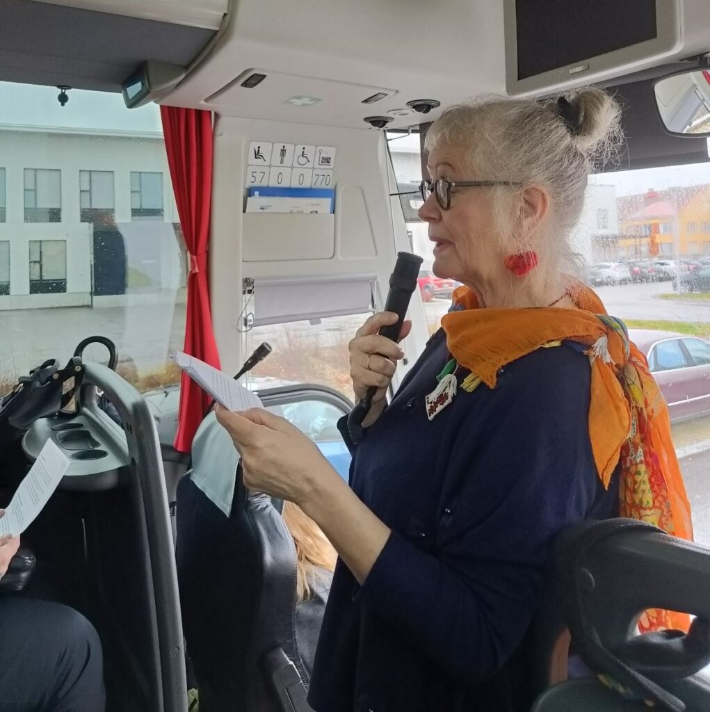 Nainen puhuu mikrofoniin. Hän seisoo linja-auton etuosassa.