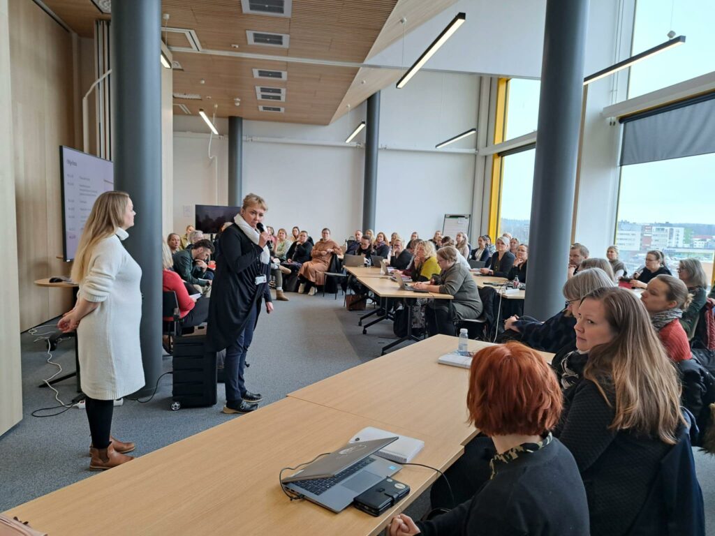 Iso joukko ihmisiä isossa kokoustilassa, ihmisten edessä pari henkilöä seisomassa, toisella kädessään mikrofoni. 
