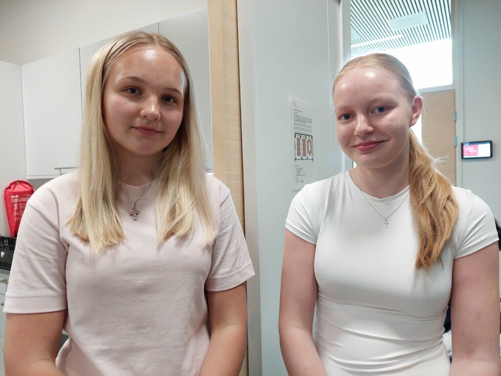 Kaksi henkilöä hymyilevät ja katsovat kameraan päin. Molemmilla vaalea t-paita yllään.