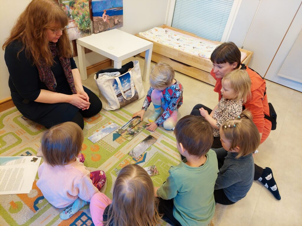Lapset istuvat aikuisen kanssa lattialla ja kokoavat palapeliä, joka kuvaa maisemataulua.