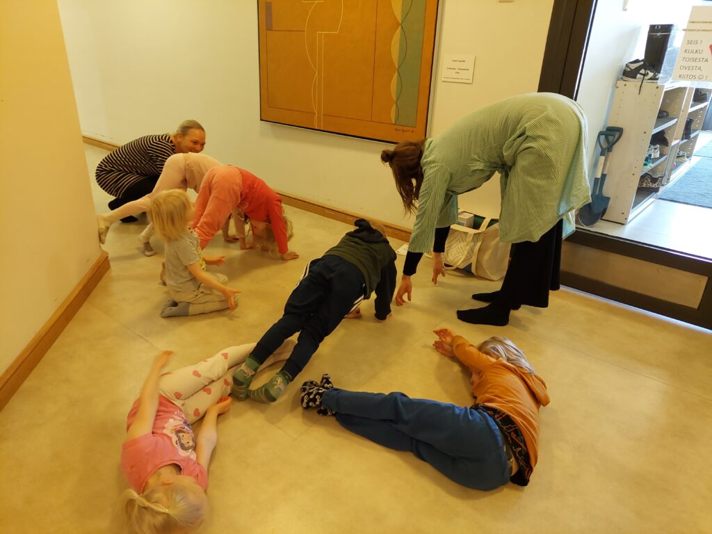 Päiväkodin käytävän seinällä on oranssisävyinen taulu, ja lapset matkivat eri asennoin taulussa olevia muotoja.