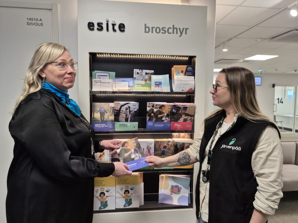 Kaksi henkilöä seisovat esiteseinämän vieressä. He pitelevät yhtä esitettä käsissään.