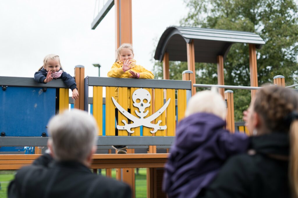 Kaksi lasta leikkivät leikkipuiston kiipeilytelineessä, etualalla aikuiset katsovat selät kameraan päin kohti lapsia.