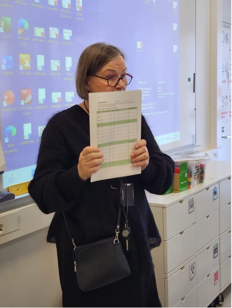 Opettaja seisoo luokan edessä ja näyttää paperilomaketta.