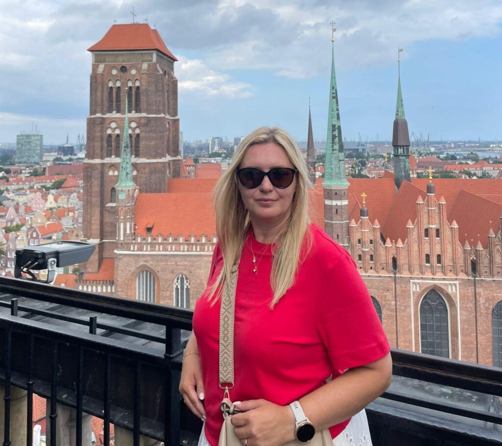 Vaaleapitkähiuksinen nainen, jolla on aurinkolasit päässä ja päällään punainen pusero. Taustalla näkyy kaupunkimaisemaa, ja kirkon torneja.