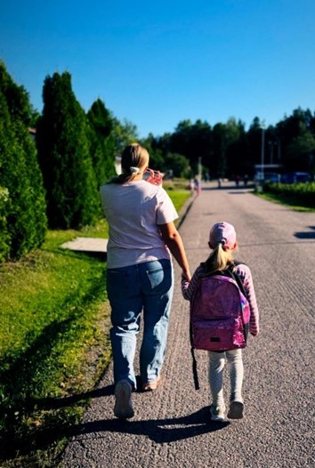 Vanhempi ja lapsi kävelevät käsi kädessä tiellä. Lapsella on reppu selässä.