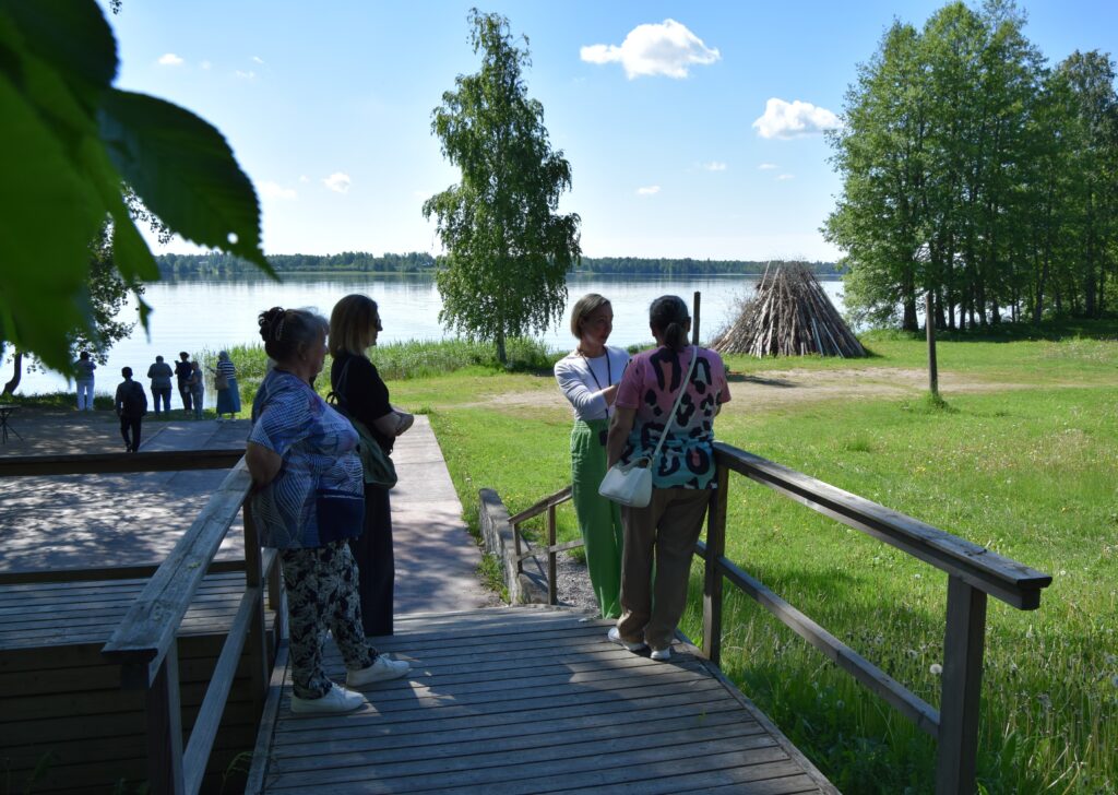Neljä ihmistä seisoo puusillalla ja katsovat valmiiksi kasattua juhannuskokkoa. Taustalla on järvi.