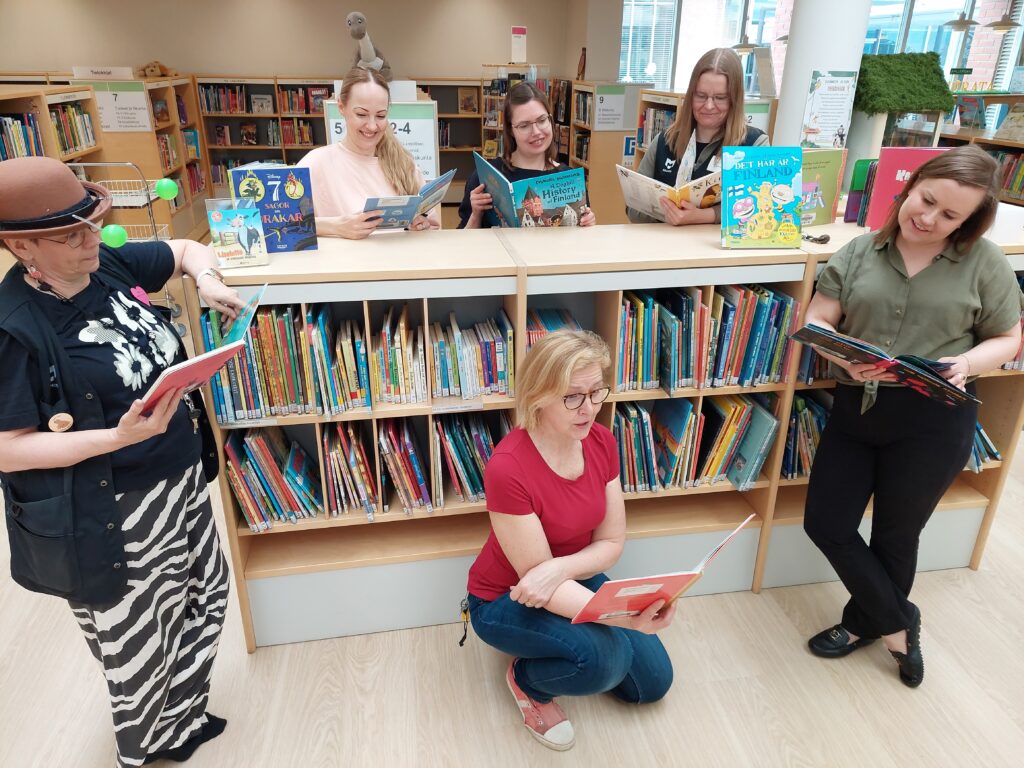 Kirjahyllyn ympärillä henkilöitä, joilla kaikilla on kirja kädessä ja he lukevat kirjaa.
