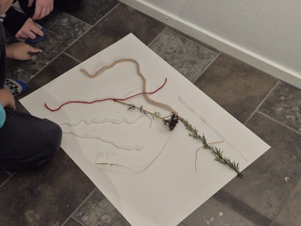 Paperilla lattialla lasten tekemää nykytaidetta. Materiaalina on käytetty luonnonmateriaalia ja lankaa.