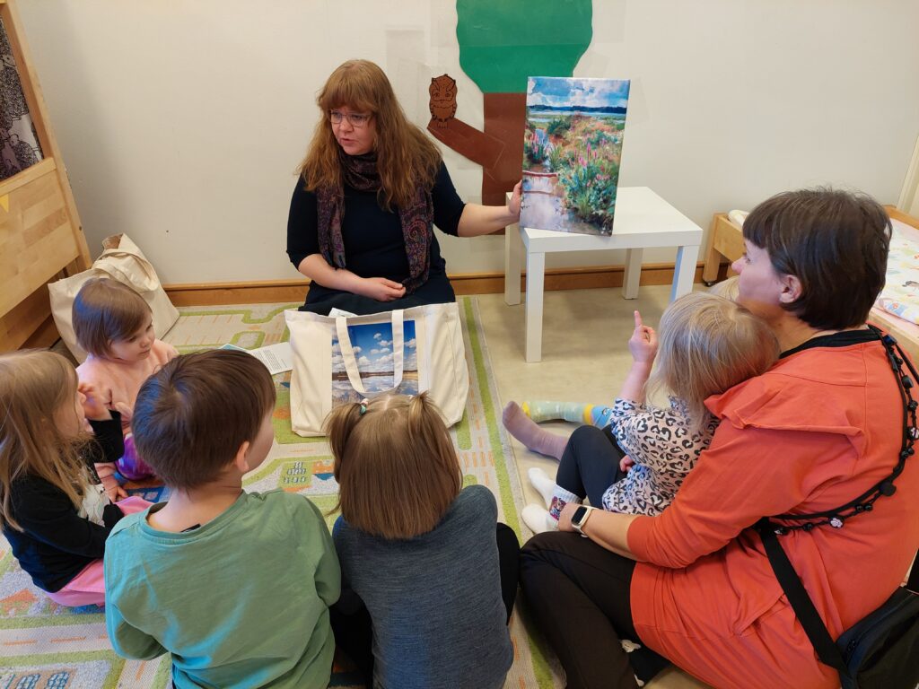 Nainen näyttää maisemataulua ympärillä oleville lapsille. Taulu esittää kukkivaa kesää.