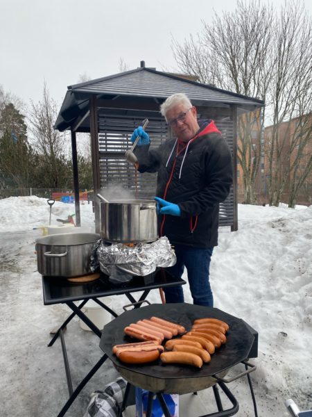 Henkilö seisoo ulkona talvisena päivänä suuren kattilan vieressä, edessään isossa valurautapannussa grillin päällä makkaroita.