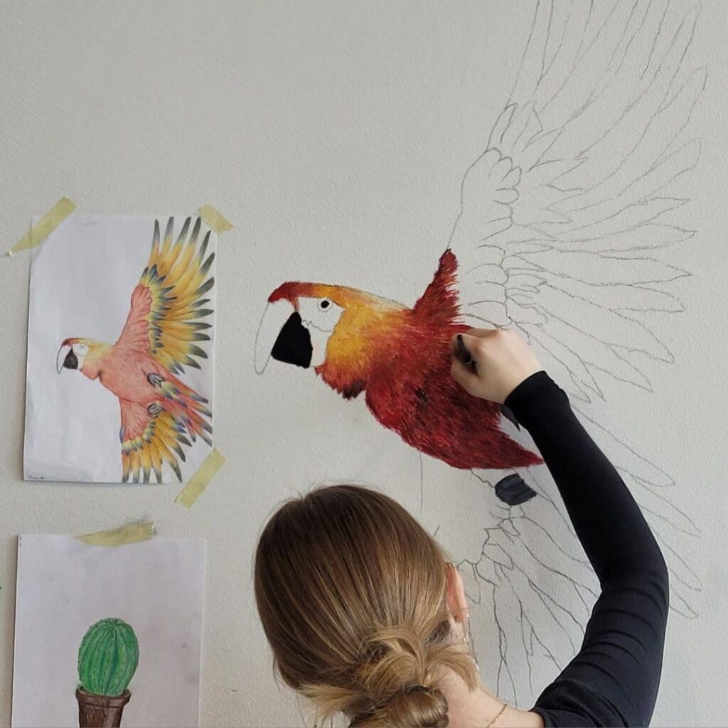 Nuori maalaamassa seinälle värikästä papukaijaa pienemmästä mallista.