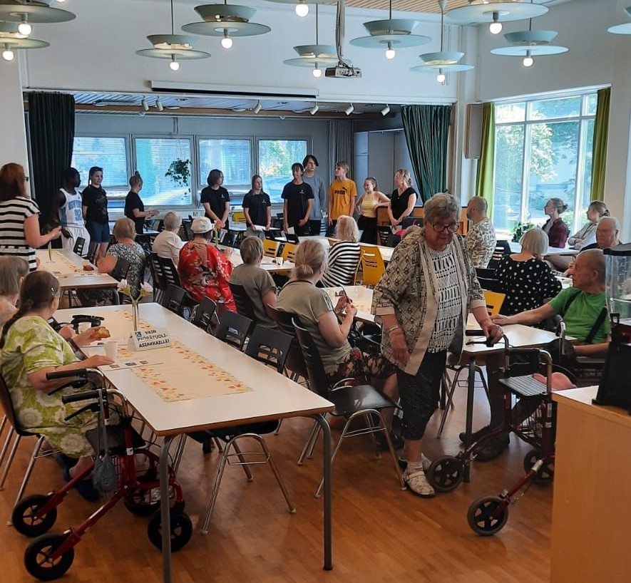 Ikäihmisiä kahvittelemassa ruokasalissa. Taustalla nuoria henkilöitä järjestämässä ohjelmaa.