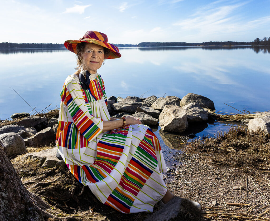Tuula Siltasari-Peltonen istuu kivellä sinisen järven rannalla yllään värikäs mekko ja hattu. Kuva: Sami Lamberg/Kapina Oy