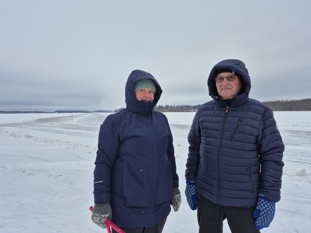Nainen ja mies sinisissä hupullisissa takeissa seisovat järven jäällä. Naisella on kädessään luistinsuojat. Takana näkyy jäällä olevaa luistinrataa.